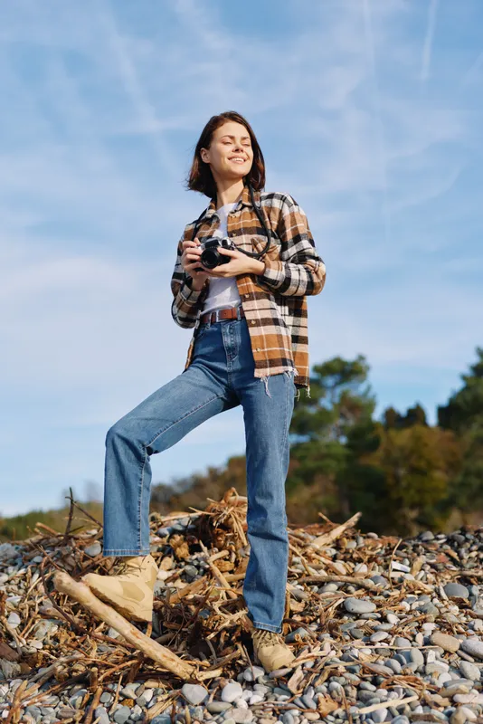 Woman photographer on a rocky shore, holding a camera, wearing plaid shirt and jeans, under bright blue sky, capturing nature and outdoor exploration.