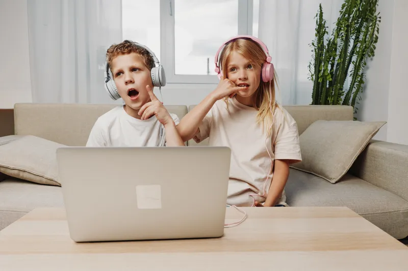 Two children wearing headphones watch a laptop screen with surprise and curiosity in a cozy living room. Concept of digital entertainment, online learning, and family time.
