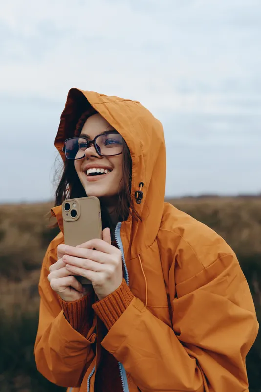 Woman outdoors in orange hooded jacket, smiling while holding smartphone, relaxed lifestyle moment, soft breeze, overcast sky, distant fields background, autumn air, nature colors