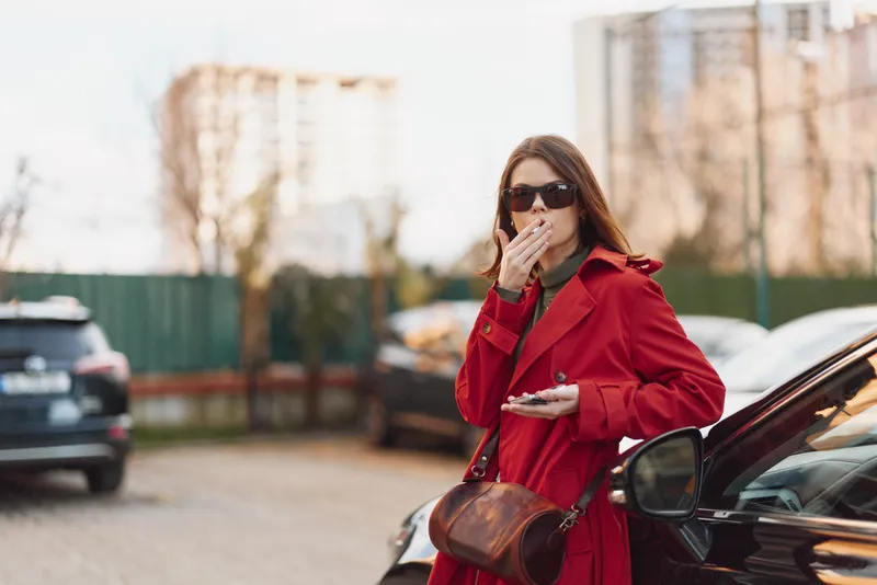 Cheerful woman outdoors in a red coat, sunglasses, eating, standing on a city street, cars in the background, daylight glow, gentle breeze, casual fashion, urban lifestyle, vivid colors