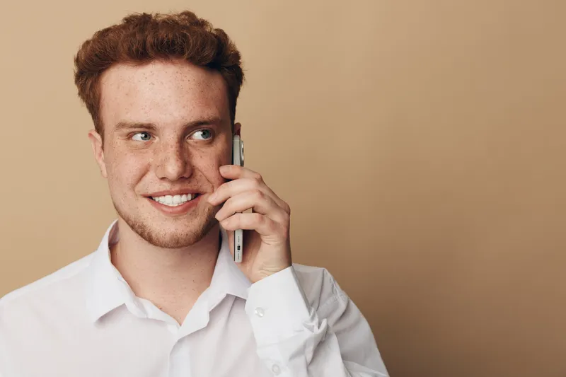 Smiling young man with curly red hair wearing white shirt talks on smartphone against beige background. Concept of communication, technology, casual lifestyle, and positive emotions.