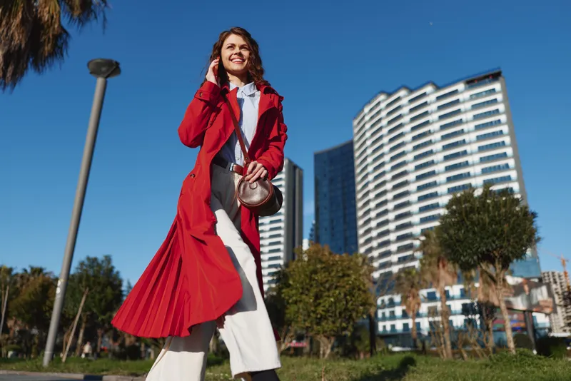 Young woman in red coat walking outdoors in urban park, smiling, sunny day, modern buildings background, casual style, confident posture