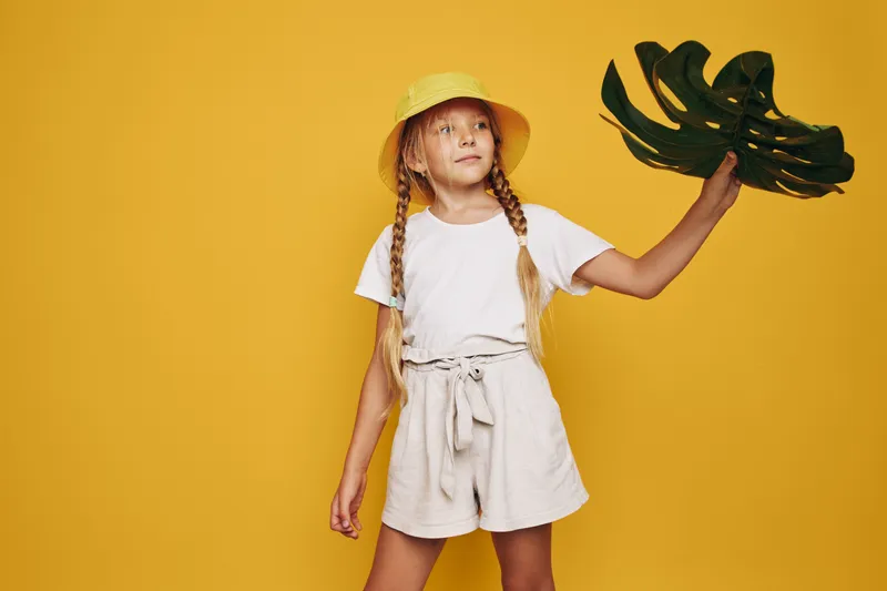 Girl in beige shorts and white t-shirt holds large green leaf against yellow background. She wears a yellow hat and looks confidently to the side. Bright studio portrait.