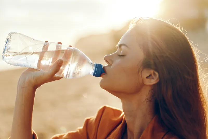 A young woman drinks water from a plastic bottle outdoors during sunset. The warm sunlight highlights her face and hair in a natural environment. Concept of hydration and refreshment.