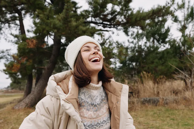 Woman enjoying an outdoor moment in a park, laughing, wearing a winter coat, beanie and sweater, with trees and open field in the background, conveying a carefree, candid lifestyle portrait for