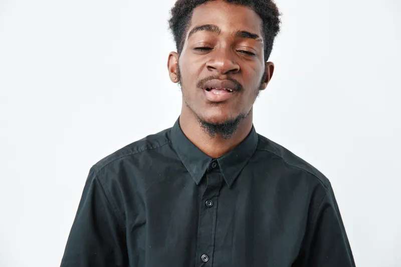 Young black man with dark skin wearing black shirt posing with eyes closed and relaxed expression on white background in studio portrait.