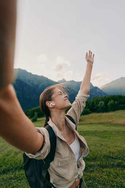 Young brunette woman with a backpack raises her arm in a sunlit field with distant mountains behind, capturing outdoorsy adventure and hopeful freedom