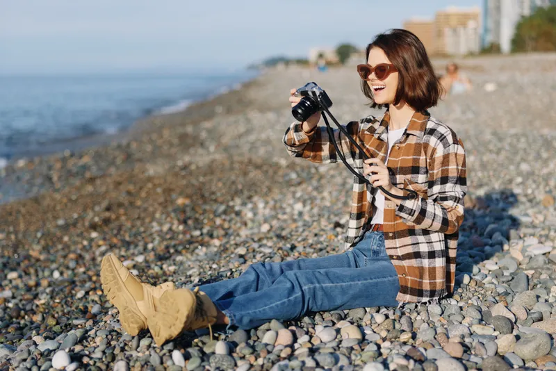 Photographer at the beach capturing scenes with a camera, casual outfit plaid shirt and jeans, sunny day on a pebbled shore, relaxed outdoor lifestyle photoshoot and creative moment