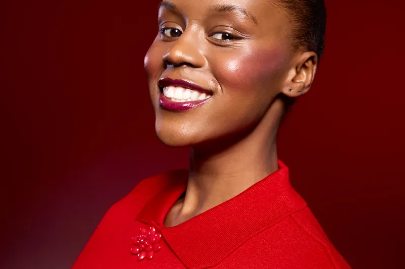 Smiling black woman with dark skin wearing a red outfit poses confidently on a red background. Close-up portrait showcasing beauty, fashion, and positive mood in studio lighting.