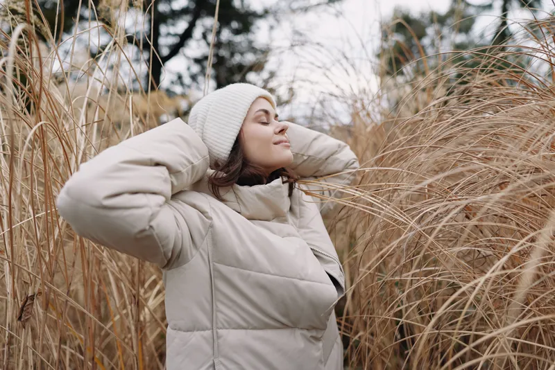 Woman in a light puffer coat enjoys outdoor calm among tall grasses, winter breeze, and soft natural light, portraying a serene fashion moment in nature and quiet mood.