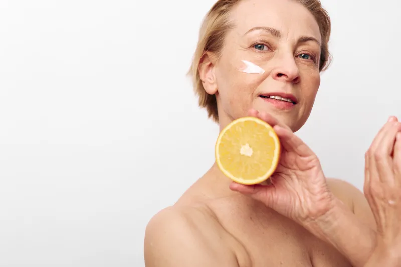 Mature woman with short hair holds half an orange near face with cream on cheek. Skincare and natural beauty concept with fresh citrus fruit in bright minimalist studio setting.