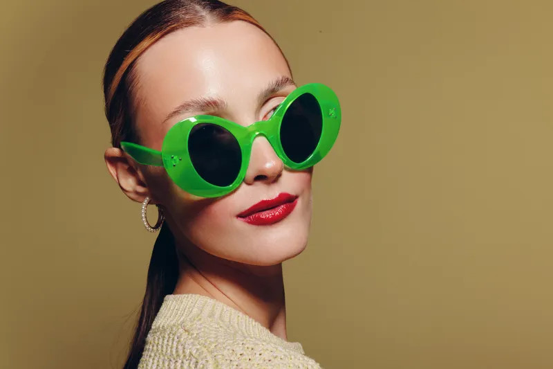 Stylish woman with green sunglasses and red lipstick poses confidently against beige background. Concept of fashion, beauty, and modern accessories in studio lighting.