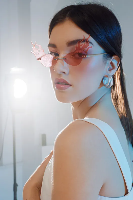 Young woman with long dark hair wearing butterfly-shaped pink sunglasses and hoop earrings poses in a white top. Soft lighting highlights her glowing skin and beauty fashion style.