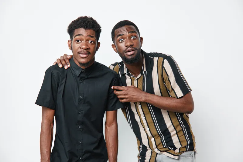 Two young black men with dark skin posing together on white background, casual clothing, one with striped shirt and the other in black shirt, friendly and surprised expressions.