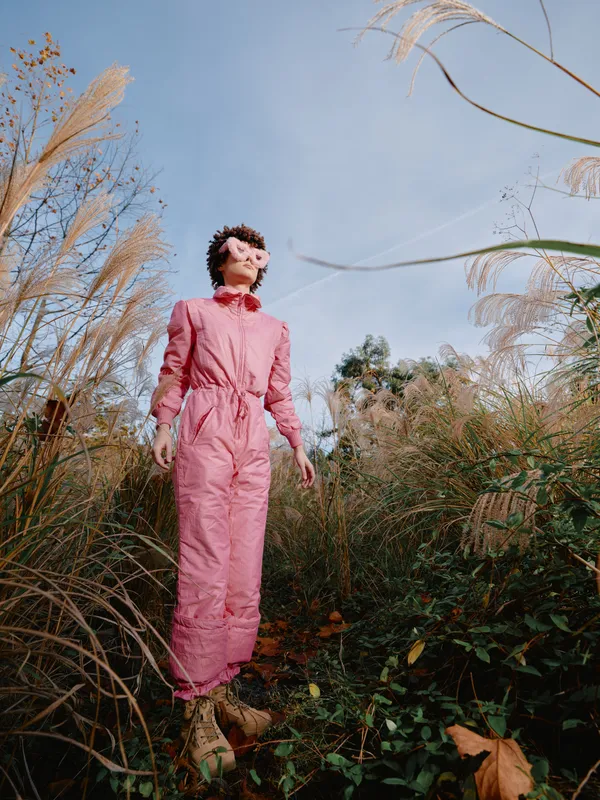Fashion model in a pink jumpsuit stands among tall grasses outdoors, sunlit scene with natural backdrop, confident pose and vibrant style in a nature setting