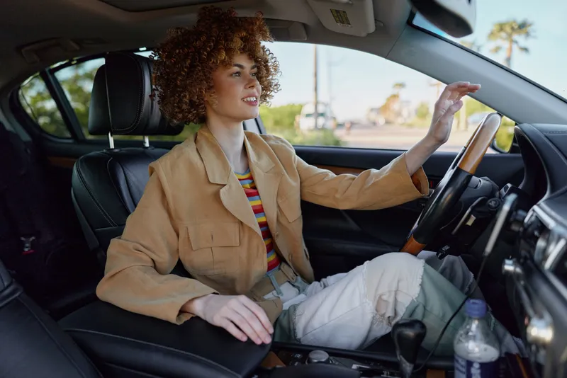 Cheerful woman driving a car, retro film color, casual outfit, sunlit interior, relaxed mood, outdoors through window, vibrant tones, candid lifestyle moment