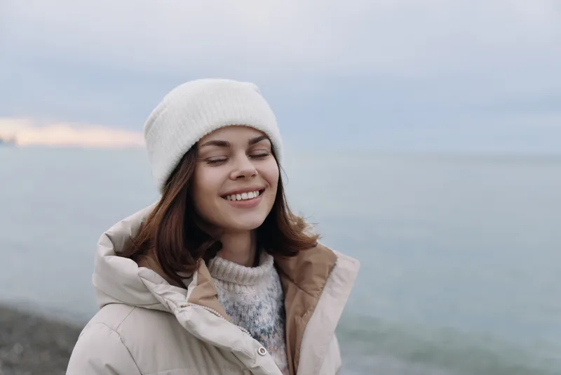 Joyful woman in a warm coat and knit hat enjoying a chilly seaside breeze, calm water behind her, soft light and relaxed mood outdoors by the coast