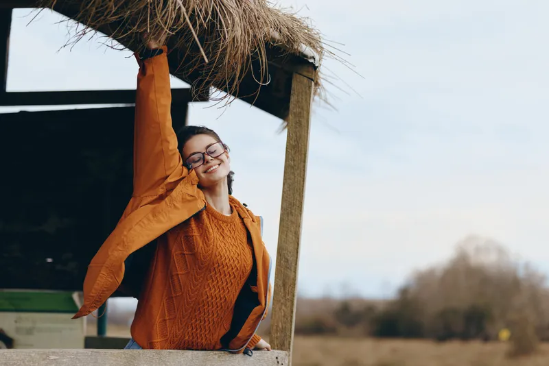 Woman lifestyle outdoors sweater glasses orange relaxation spring portrait leisure. Smiling woman in orange knit sweater and glasses, relaxing on wooden patio, daylight, blurred trees.