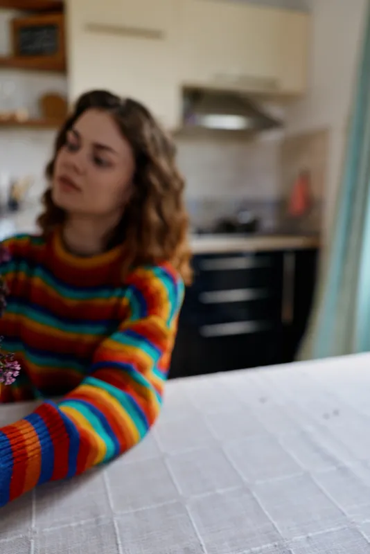 A woman in a colorful sweater leans toward the foreground in the kitchen at home, reaching across the table, showcasing casual morning dining lifestyle in a bright, intimate setting.