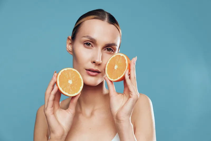 Young woman with clean skin holding two halves of an orange near her face. Blue background with soft lighting highlights natural beauty and healthy lifestyle concept.