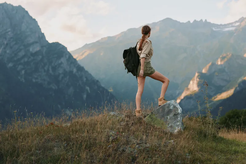 Young woman with a backpack stands on a rock overlooking a mountain landscape during a hiking trip, enjoying the outdoor adventure and natural scenery.