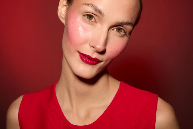 Close-up of a smiling woman with red lipstick and pink blush wearing a red sleeveless top against a red background. Concept of beauty, makeup, fashion, and confidence.