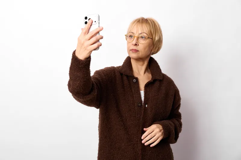 Elderly woman with glasses taking a selfie with a smartphone, indoor portrait on a white background, wearing a brown coat, soft studio light, calm friendly mood today