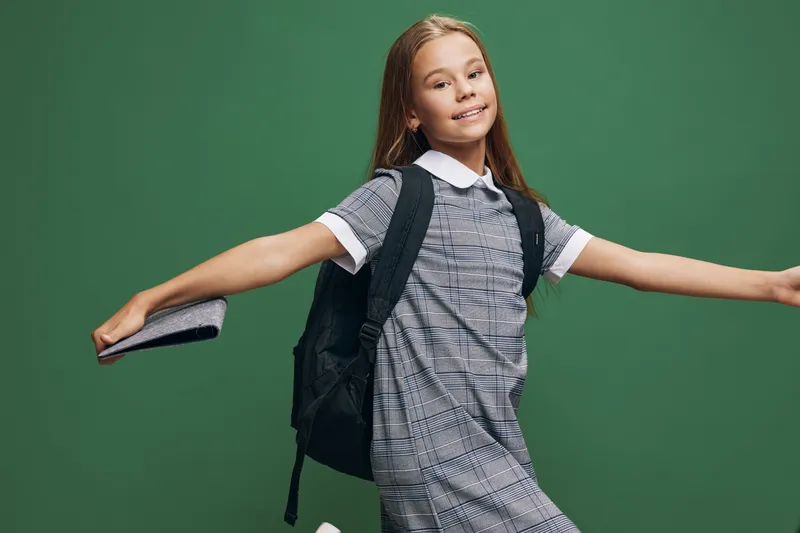 Girl with long hair wearing checkered dress and backpack smiling happily while posing with arms outstretched holding a book on green background in studio portrait.