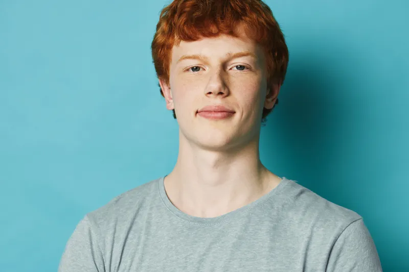 Young man with red hair wearing gray t shirt stands against blue background looking confidently at camera with slight smile on face and relaxed posture