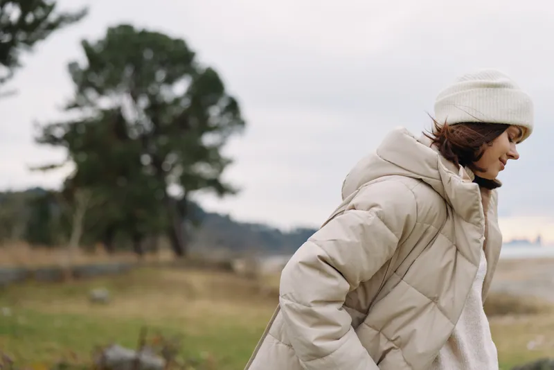 Woman in a beige puffer coat walks outdoors on a breezy day with a knit hat, in a natural setting of trees and open field, conveying seasonal fashion and calm