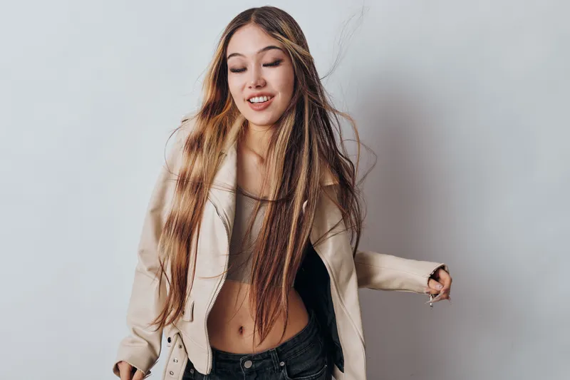 Young woman with long braided hair smiling and posing wearing light jacket and casual top against plain background. Female model with relaxed expression and stylish outfit.
