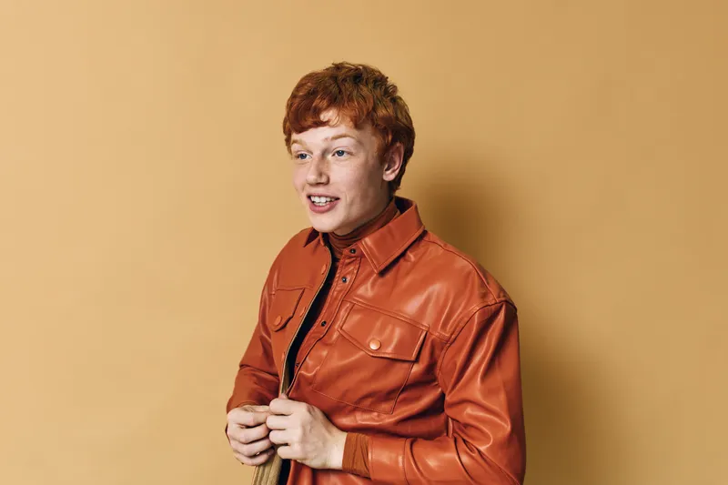 Young man with red hair smiling and wearing orange leather jacket standing against beige background. Casual style, happy expression, studio portrait of confident male.