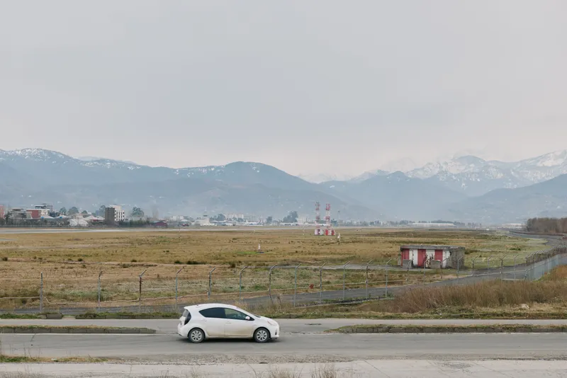White compact car parked on rural road with dry grass fields and distant snow covered mountains under overcast sky. Calm landscape with muted colors and open space.