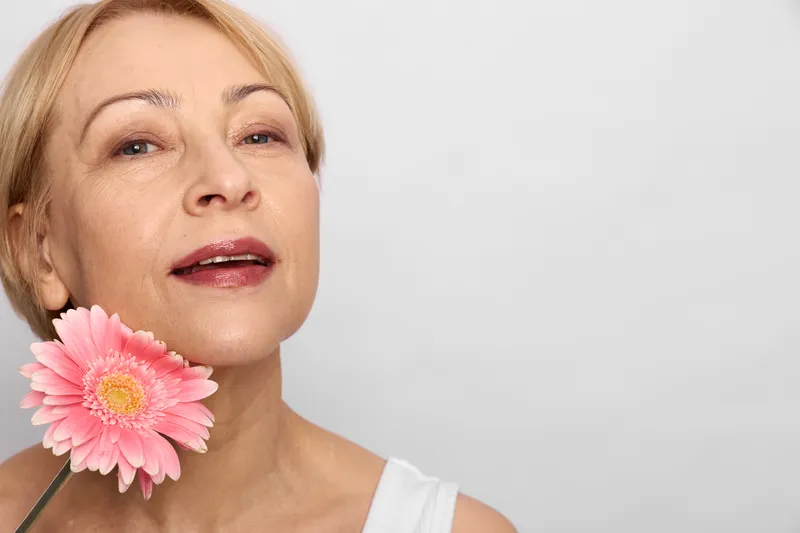 Woman portrait closeup with pink flower near face, blonde hair, gentle smile, soft studio light, beauty makeup, spring mood, calm expression, white background