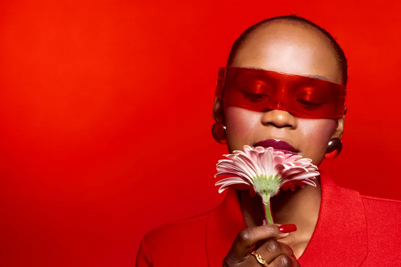 African woman with dark skin wearing red sunglasses and red blazer holds pink flower close to face on red background. Concept of fashion, beauty, elegance, and floral aesthetics.