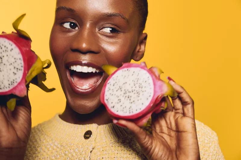 Smiling dark skinned African woman holds two halves of dragon fruit close to face. Yellow background and cozy sweater create warm joyful mood. Concept of healthy lifestyle and exotic fruit.