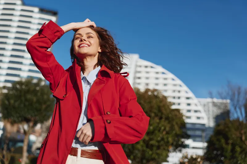 A confident young woman in a red trench smiles toward the sun in an urban square, expressing optimism and empowerment.