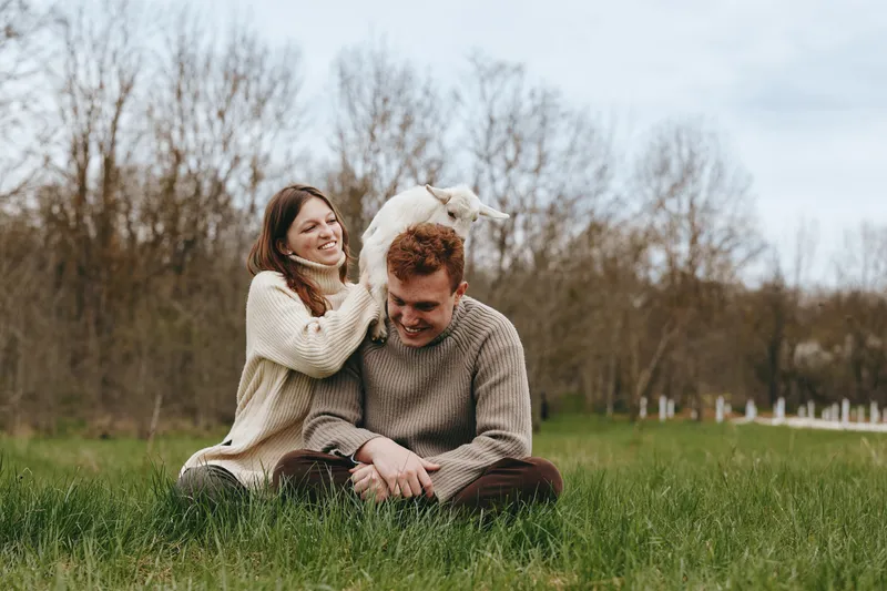 Cheerful couple outdoors, close in with a playful goat kid by their side, cozy sweaters, sunlit meadow, natural colors, relaxed posture, candid moment, nature backdrop, lifestyle portrait