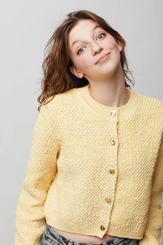 Young woman with brown hair wearing yellow knitted cardigan smiles gently looking at camera. Casual style, natural makeup, isolated on light background. Concept of happiness and youth.