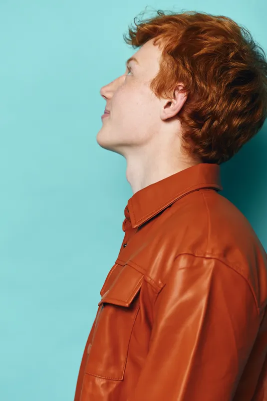 Young man with red hair wearing brown jacket posing in profile looking upwards with thoughtful expression against blue background in studio portrait of casual fashion style.