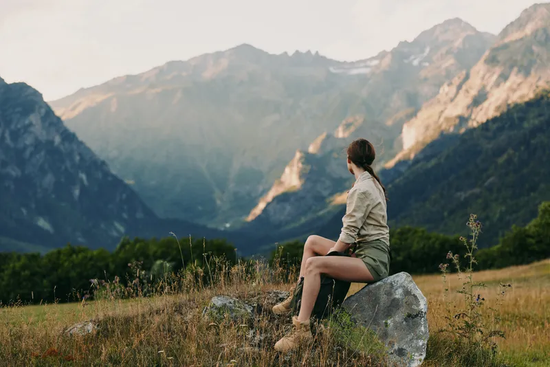 Young woman with long hair sits on a rock in a mountain meadow enjoying the scenic view of distant peaks during a calm outdoor moment.