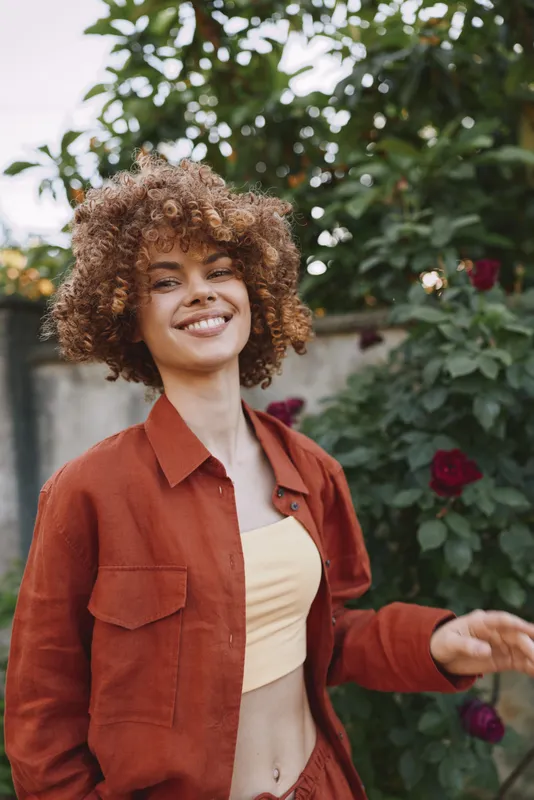 A cheerful woman with curly hair wearing an orange shirt jacket and cream top stands outdoors among blooming roses, capturing a playful mood and personal style. Concept: carefree, confidence