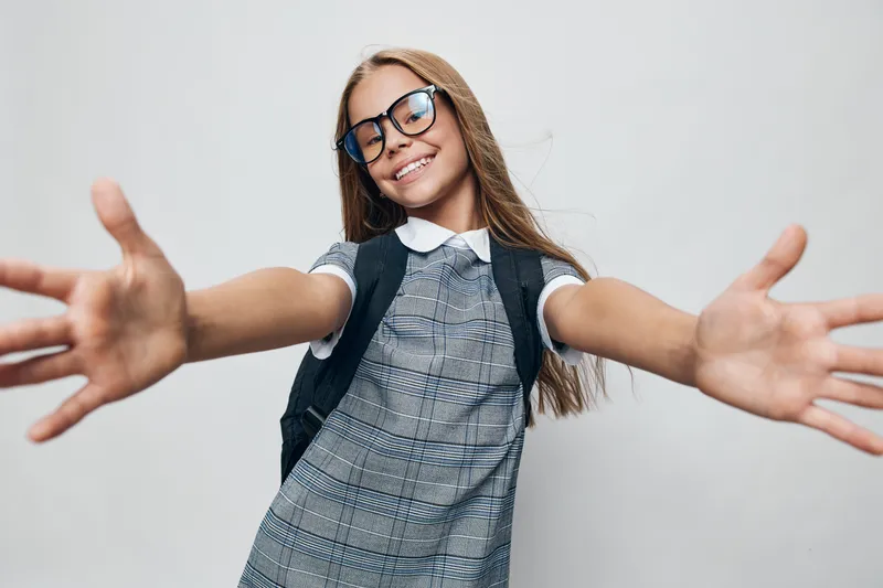 Girl with glasses and backpack smiling with arms wide open for welcome or hug gesture. Portrait of happy child in casual dress with long hair on white background.