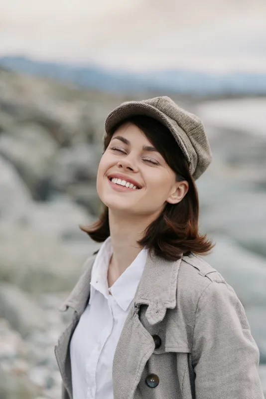Smiling young woman in casual beige jacket and hat enjoys outdoor nature moment. Soft light highlights relaxed mood and natural beauty with blurred rocky background.