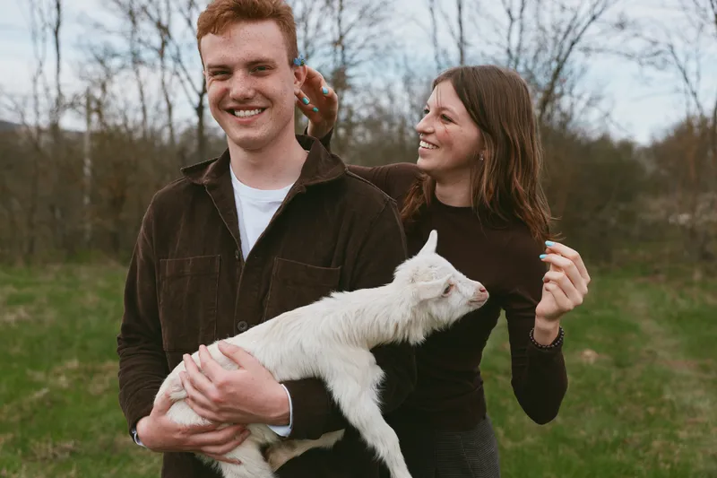Cheerful couple outdoors holding a white goat kid, smiling together, casual clothing, soft breeze, sunlit grass, natural colors, candid lifestyle moment, relaxed connection, countryside vibe