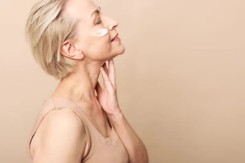 Mature woman with short blonde hair applies cream on face and neck, eyes closed, wearing beige top. Concept of skincare, antiaging, selfcare, and natural beauty on neutral background.
