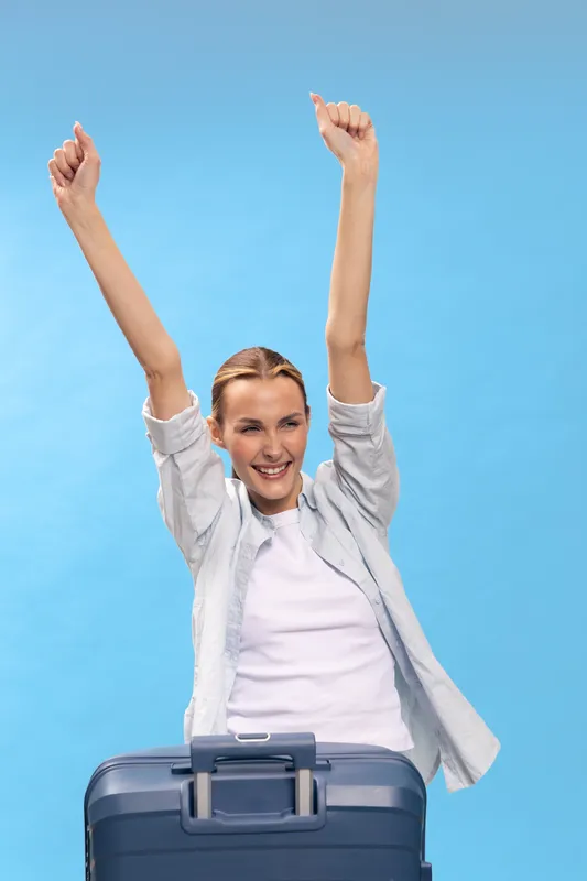 Happy woman in casual clothes raises arms in celebration behind blue suitcase on light blue background. Concept of travel, vacation, joy, freedom, and summer holiday excitement.