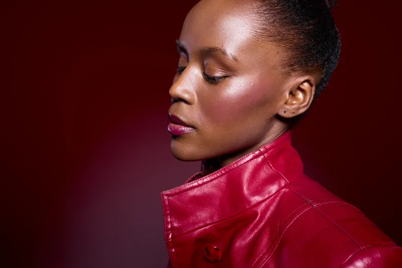 Close-up portrait of elegant African woman with dark skin in red leather jacket against dark red background. Concept of fashion, beauty, and confident female model in studio lighting.