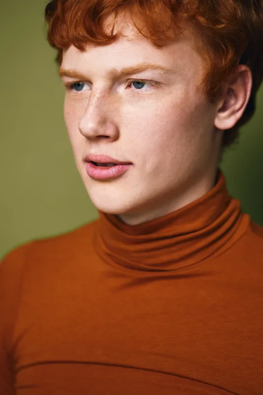 Portrait of young man with red hair and blue eyes wearing orange turtleneck sweater, looking sideways on green background with serious expression and clean skin in studio light.