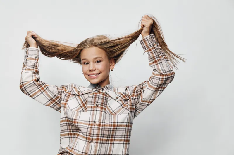 Girl smiling and holding her long hair with both hands in casual plaid shirt on light background. Child posing happily with playful expression and relaxed mood.